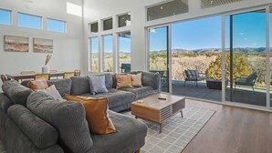 A spacious living room with a large sectional sofa, a wooden coffee table, and a dining area with a table and chairs. Large windows offer a view of the surrounding landscape. - The Grove-River Cabin -Hot Tub- Family Home Near Bryce & Zion (Hatch)