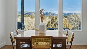 A dining table with six chairs, a vase with dried flowers, and a view of mountains through large windows. - The Grove-River Cabin -Hot Tub- Family Home Near Bryce & Zion (Hatch)