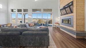 A spacious living room with a large sectional sofa, a fireplace, and large windows offering a view of the outdoors. - The Grove-River Cabin -Hot Tub- Family Home Near Bryce & Zion (Hatch)