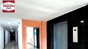 Un pasillo de hotel con diseño moderno, que presenta un suelo de baldosas de mármol, un corredor central con puertas a ambos lados y una pantalla digital en la pared.