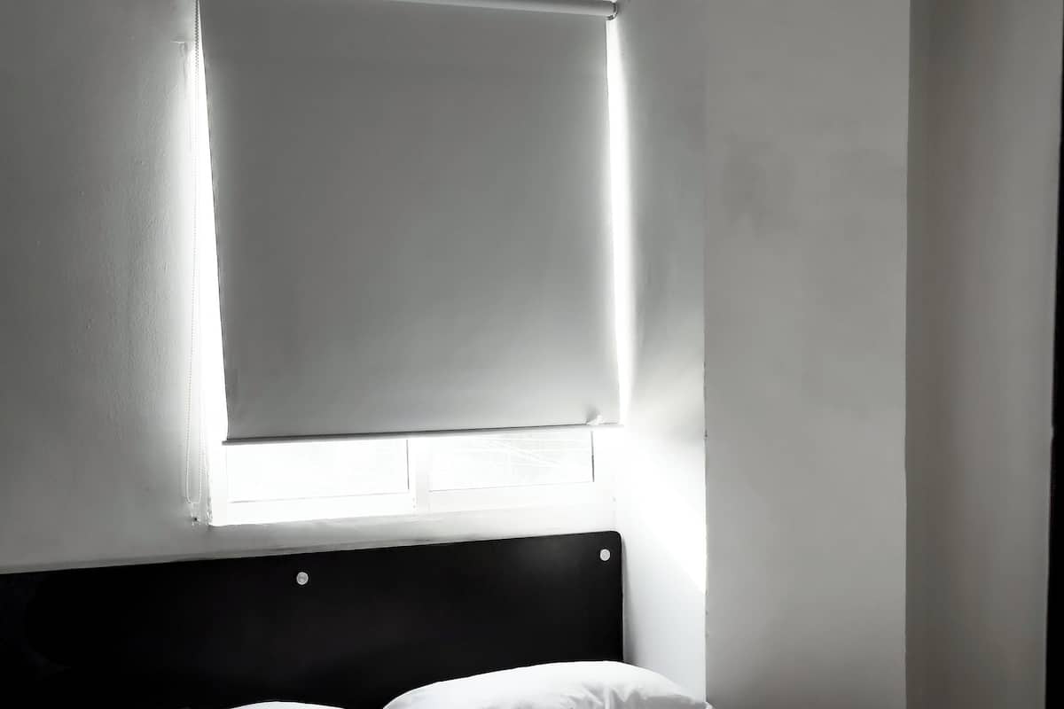 Una cama bien tendida con sábanas blancas en una habitación con paredes blancas y una ventana con persianas.