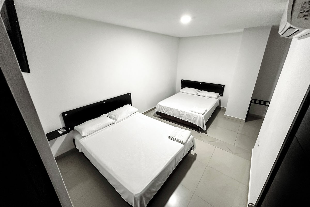 Habitación con dos camas, cada una con sábanas blancas y cabeceros oscuros. Las paredes son blancas y el piso, de baldosas.