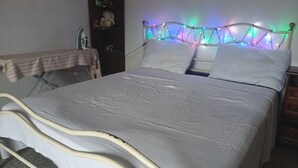 A bed with a white mattress and headboard, a bedside table with a striped cloth, and a wall-mounted shelf with various items.