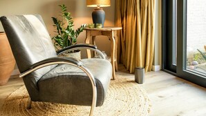 A modern living room with a leather armchair, a wooden side table with a lamp, a potted plant, and a view through a large window. - Komfortabel Eingerichtetes Ferienhaus mit Herrlichem Sonnige Terrasse (Heiloo)