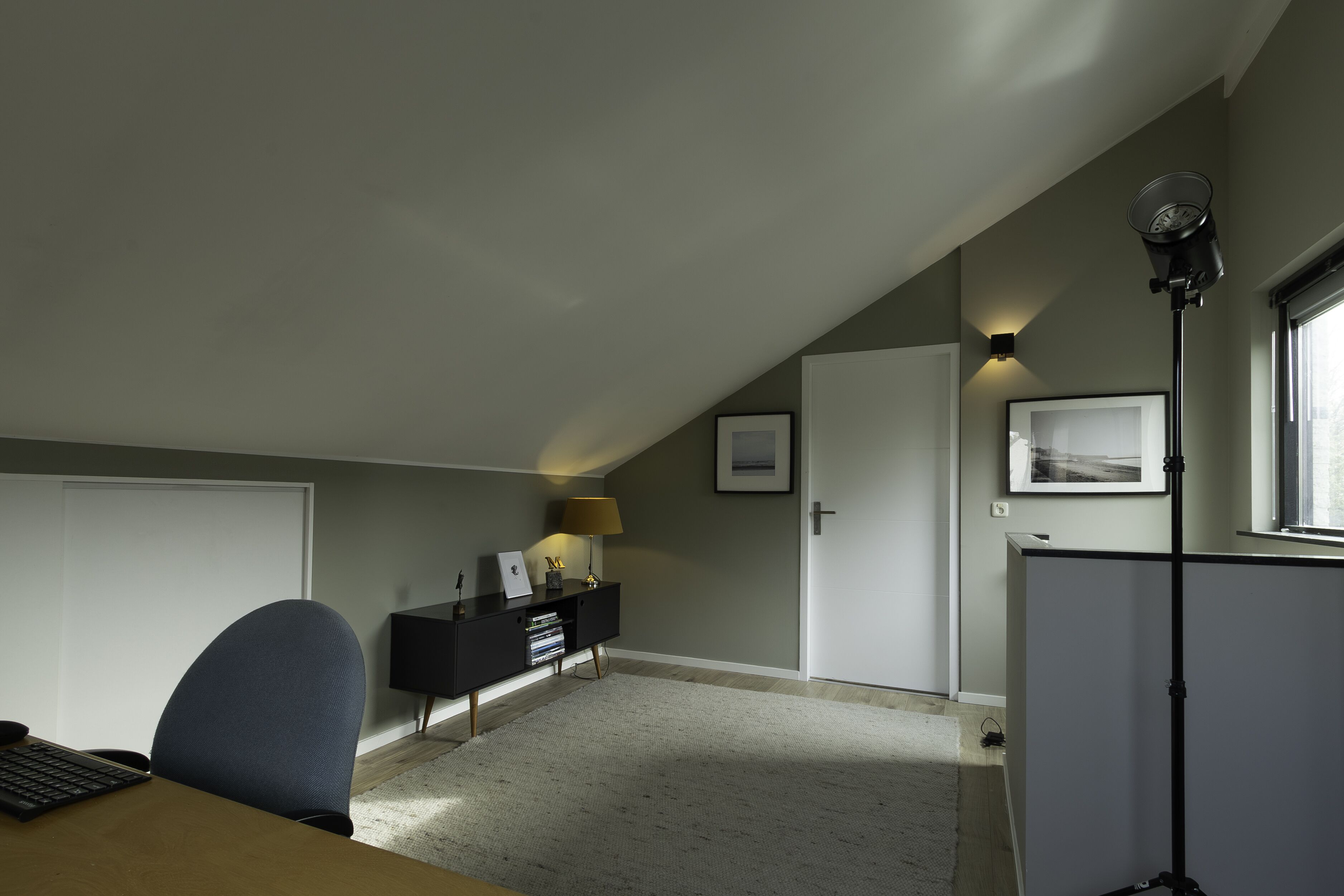 Een kamer met een bureau, stoel, zwarte console, lamp, lijst met afbeelding en een staande lamp.