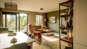 A cozy living room with a sofa, coffee table, and dining table. There is a bookshelf and a window with curtains. - Komfortabel Eingerichtetes Ferienhaus mit Herrlichem Sonnige Terrasse (Heiloo)