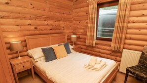 A wooden cabin bedroom with a bed, nightstands, a chair, and a window with curtains. - 24 Lakeside Drive (Morpeth)