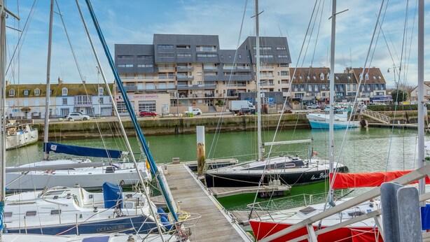 A marina with several boats docked, a wooden pier, and a row of residential buildings in the background. - Un Balcon sur le Port - Place de parking (Courseulles-sur-Mer)