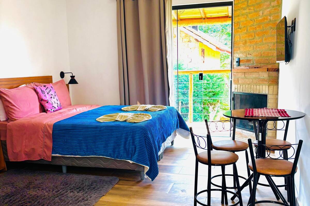 Een slaapkamer met een bed, een klein tafeltje met stoelen, een open haard en uitzicht op groen.