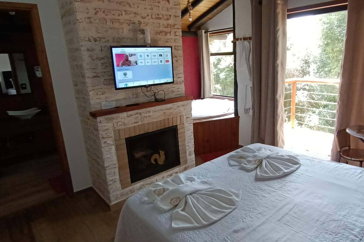 Een hotelkamer met een schouw van baksteen, een bed met een witte spreidek en decoratieve kussens, een aan de muur bevestigde televisie, en een balkon met uitzicht op bomen.