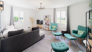 Uma sala de estar moderna com um sofá cinza, poltronas verde-água e pufes. Uma estante e uma televisão de tela plana são visíveis ao fundo.