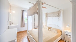 Quarto com cama de dossel, mesa de cabeceira com abajur, mesa de apoio e janela com cortinas.
