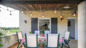 Um pátio externo com mesa posta para quatro pessoas, rodeado por cadeiras, sob um teto de madeira com luminárias pendentes.