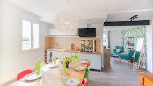 Uma cozinha moderna com televisão, uma sala de jantar com mesa posta para quatro pessoas e uma sala de estar com cadeiras cor turquesa e um quadro.