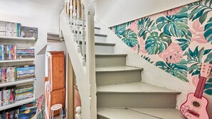Un escalier orné d’un mur décoré de motifs de feuilles tropicales, une guitare rose et une bibliothèque remplie de livres.