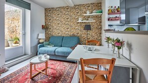 Un espace de vie compact avec un mur en pierre, un canapé, une table à manger avec des chaises, un petit tapis et une fenêtre donnant sur un mur en pierre à l’extérieur.