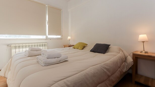 A neatly made bed with a white duvet, two pillows, and a stack of white towels. There are two bedside tables with lamps, one on each side of the bed. - Mystico (Ushuaia)