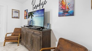 A living room with a television on a wooden cabinet, two brown chairs, and a wall with a 'Paradise' sign and two framed pictures. - Prime Downtown Key West Spot - The Cozy Little Cottage! (Key West)