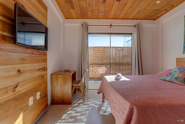 A bedroom with a wooden ceiling, a bed with a pink bedspread, a desk with a chair, and a TV mounted on the wall. - Mar e rio Orla Itacaré - Casa Flor (Itacaré)