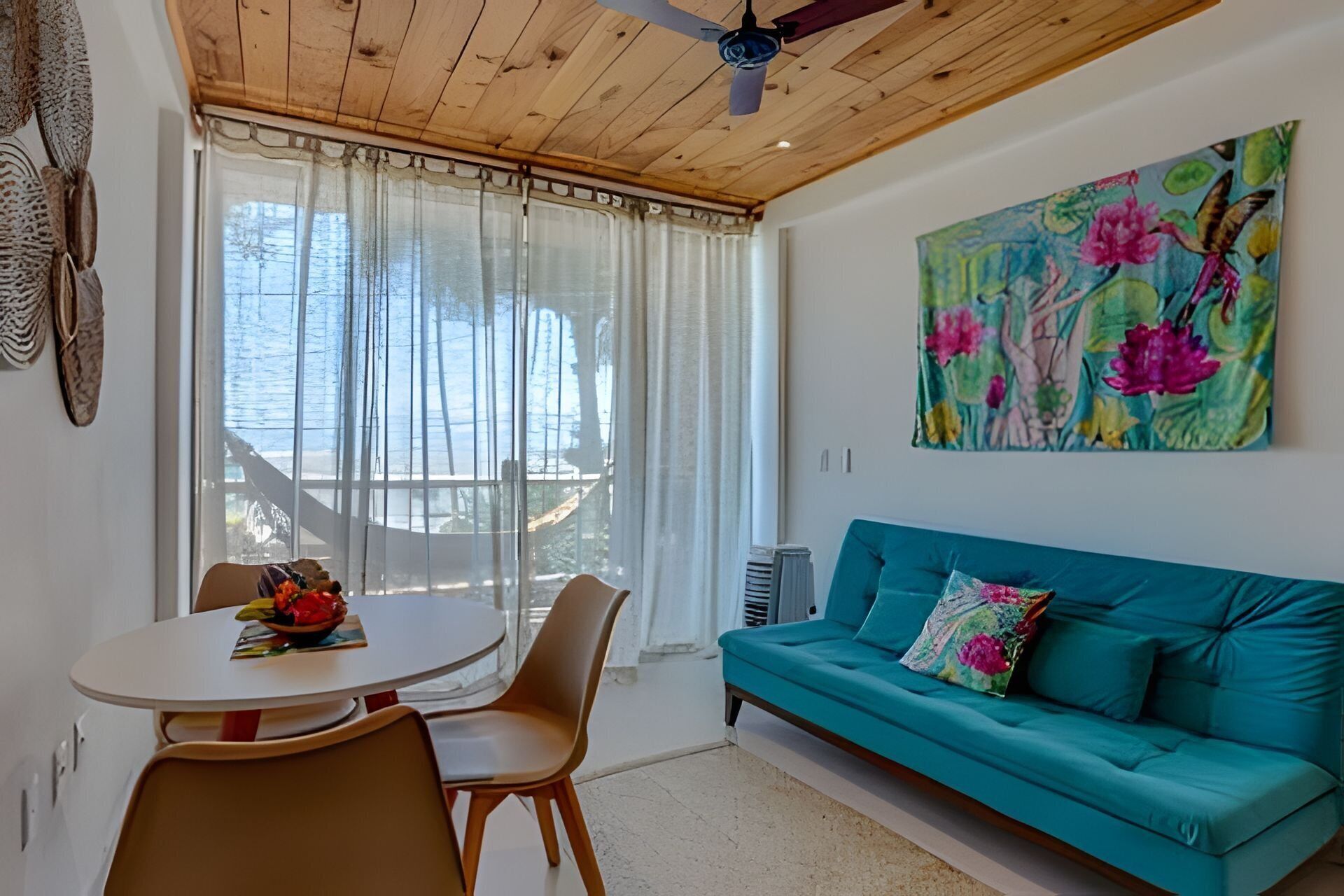 Ein Zimmer mit Holzboden, einem salbeigrünem Sofa mit einem Blumenkissen, einem runden Tisch mit Stühlen und einem großen Fenster mit durchsichtigen Vorhängen.