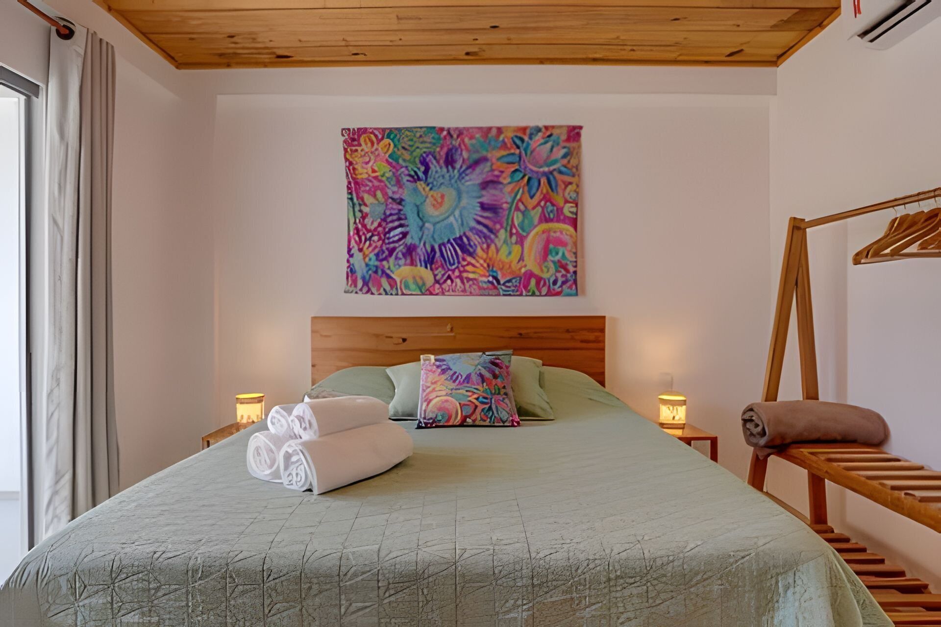 A bedroom with a wooden headboard, a colorful abstract painting above the bed, a green bedspread, and a wooden chair by the window.