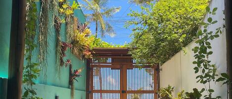 A narrow pathway leading to a gated entrance with a bamboo roof, surrounded by lush greenery and a clear blue sky.