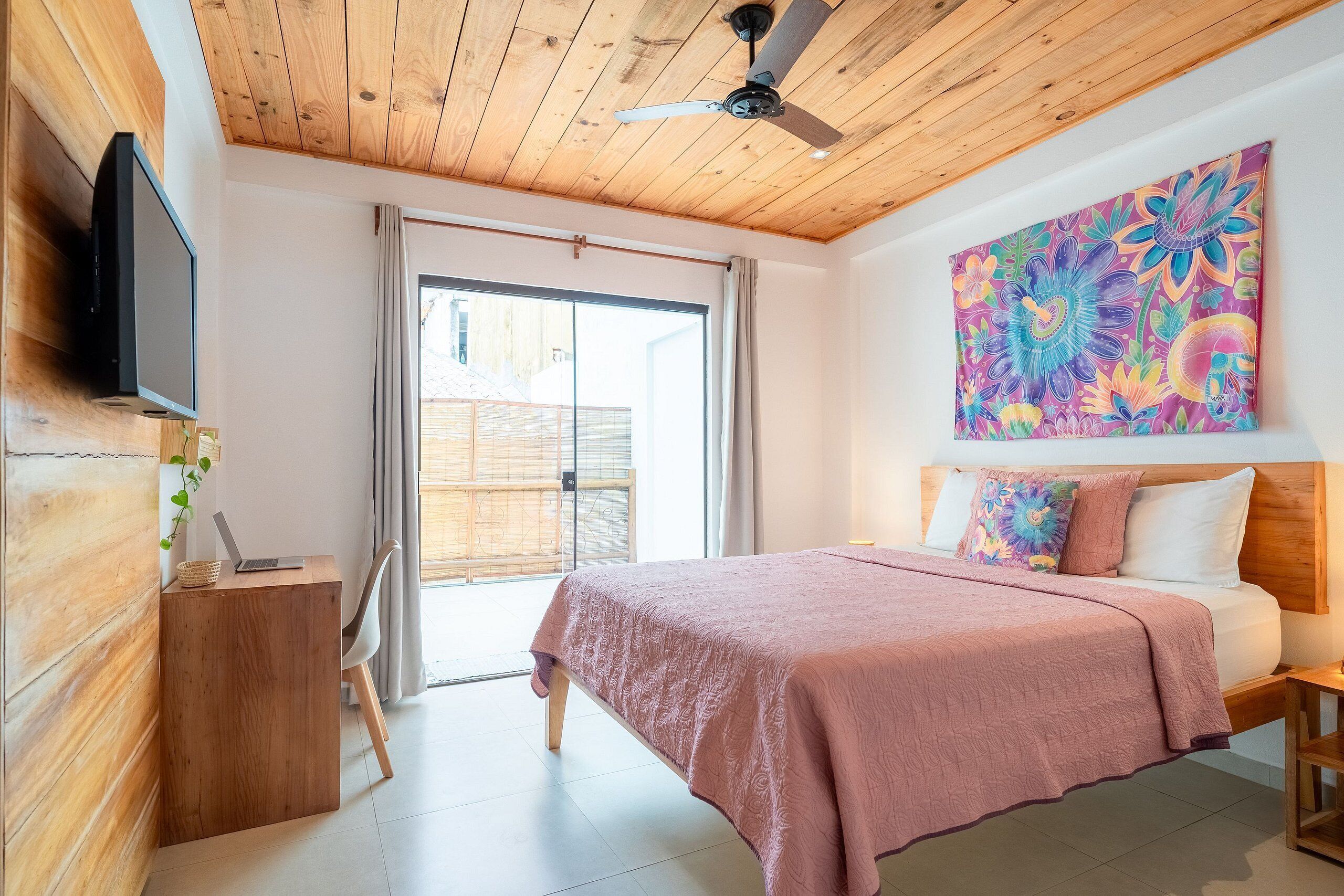 A bedroom with a wooden ceiling, a bed with a pink bedspread, a ceiling fan, a TV on the wall, and a large floral painting.