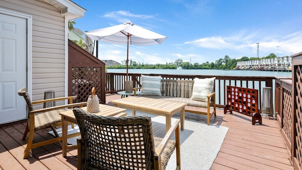 A wooden deck with outdoor furniture, a white umbrella, and a view of a body of water and buildings in the background.