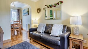 Un soggiorno con divano in pelle, un tavolo da pranzo con sedie, un lampadario, uno specchio decorato con una ghirlanda floreale e una lampada su un tavolino in legno.
