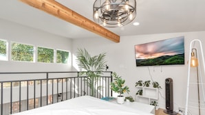 Ein Schlafzimmer mit einem großen Bett, einem Balkon, einem Topf mit Pflanze, einem an der Wand montierten Fernseher und einem Standventilator.