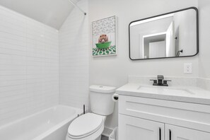 Ein modernes Badezimmer mit weißen Fliesen, einer Badewanne, einer Toilette, einem Waschbecken mit Spiegel und einem gerahmten Bild an der Wand.