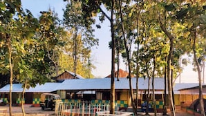 A rustic outdoor area with a dirt ground, a small auto-rickshaw, and a row of colorful buildings with corrugated metal roofs. - Resort Ichche Kuthi by Urban Express (Bangaon)