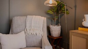 Una habitación acogedora con un sillón gris, una lámpara de pie, una planta en maceta y una pequeña mesa de madera con una taza blanca y libros.