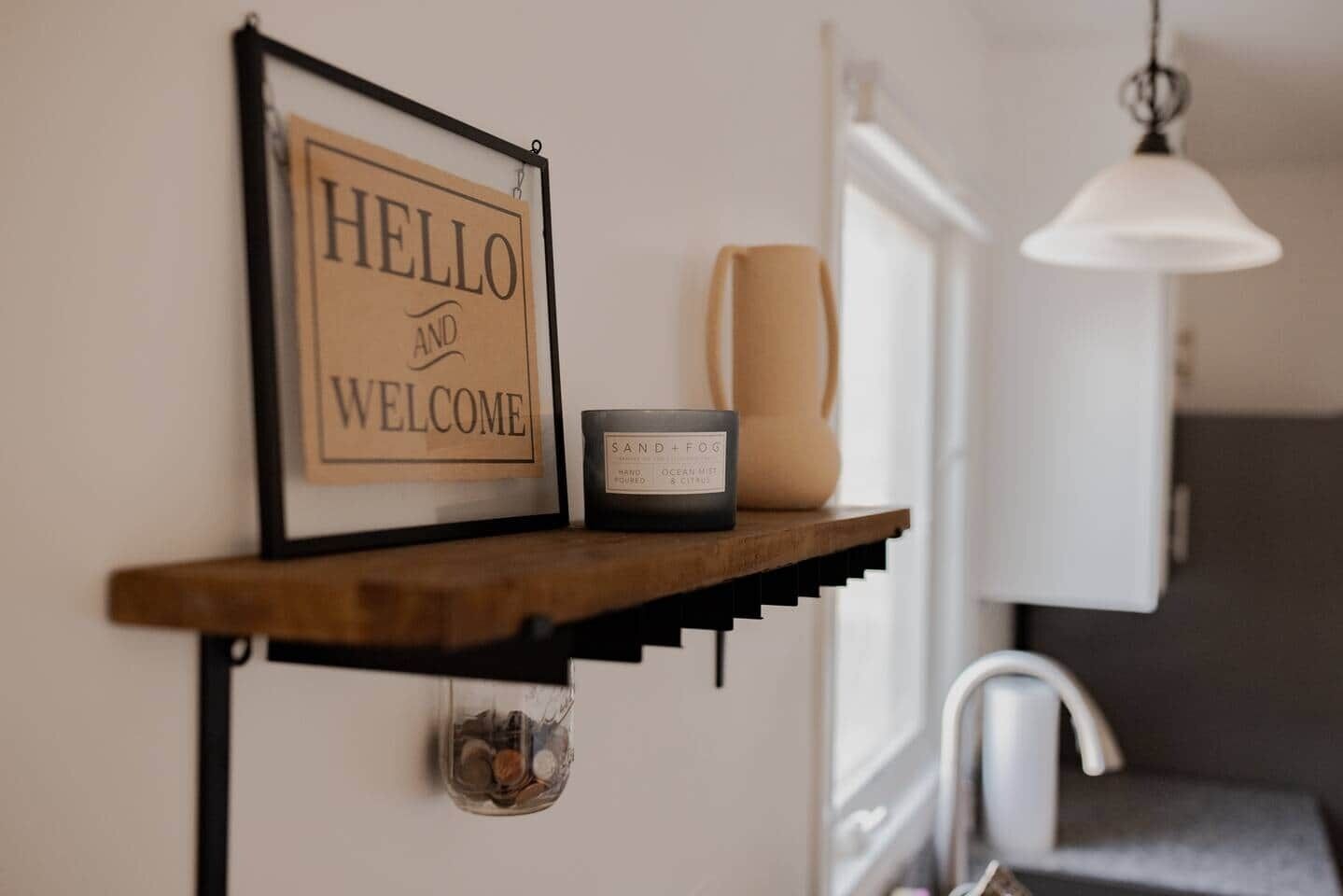 A wall-mounted shelf with a 'HELLO AND WELCOME' sign, a candle labeled 'SAND+FOG,' and a small container of coins.