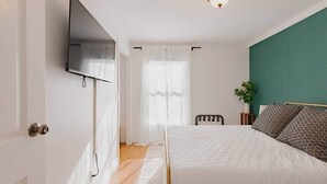 Un dormitorio con una cama, un televisor montado en la pared, una silla, una mesita de noche con una planta y una ventana con cortinas translúcidas.