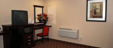 A hotel room with a desk, a television, a chair, a vase with flowers, a framed picture on the wall, and an air conditioning unit.