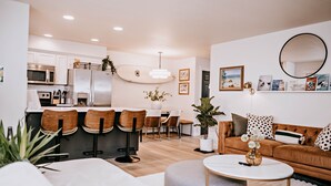 A modern kitchen with stainless steel appliances, a dining area with wooden chairs, and a living room with a brown sofa and a round coffee table.