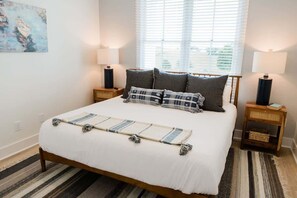 A neatly made bed with a white duvet and patterned pillows, flanked by two bedside tables with lamps, in a room with a striped rug and a window with blinds.