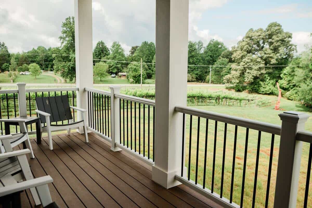 A covered porch with white railings and wooden flooring, overlooking a grassy area with trees and a vineyard.
