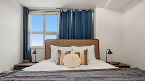 Een net opgemaakt bed met een houten hoofdeinde, twee nachtkastjes met lampen, een raam met blauwe gordijnen en een uitzicht op de lucht.