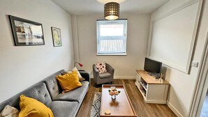 Ein modernes Wohnzimmer mit einem grauen Sofa, einem kleinen Sessel, einem Holztisch, einem Fernseher auf einem Schrank und einem Fenster mit Jalousien.