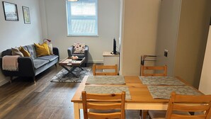 Ein Wohnzimmer mit einer Couch, einem Couchtisch, einem Esstisch mit Stühlen, einem Fernseher und einem Fenster mit Jalousien.