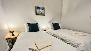 Ein Hotelzimmer mit zwei Betten, weißer Bettwäsche, dunklen Kopfkissen, einem gerahmten Bild an der Wand und einem gefalteten Handtuch auf dem Bett.