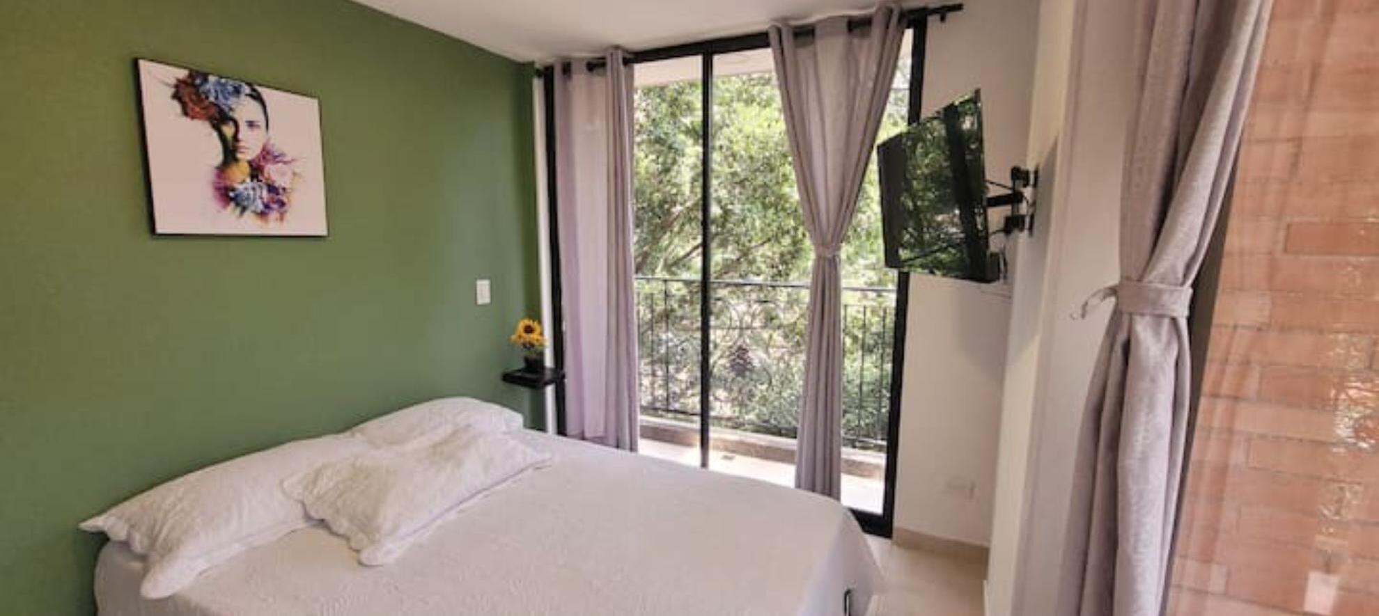 A bedroom with a bed, a painting on the wall, a TV mounted on the wall, and a window with curtains.