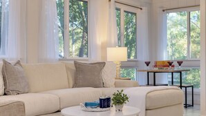 A living room with a sofa, a coffee table, and a lamp. There are windows with curtains, a small table with board games, and a plant on the coffee table. - Conch Cottage by Avantstay Walk to Beach, Deck, Fireplace, Outdoor Dining (Isle of Palms)
