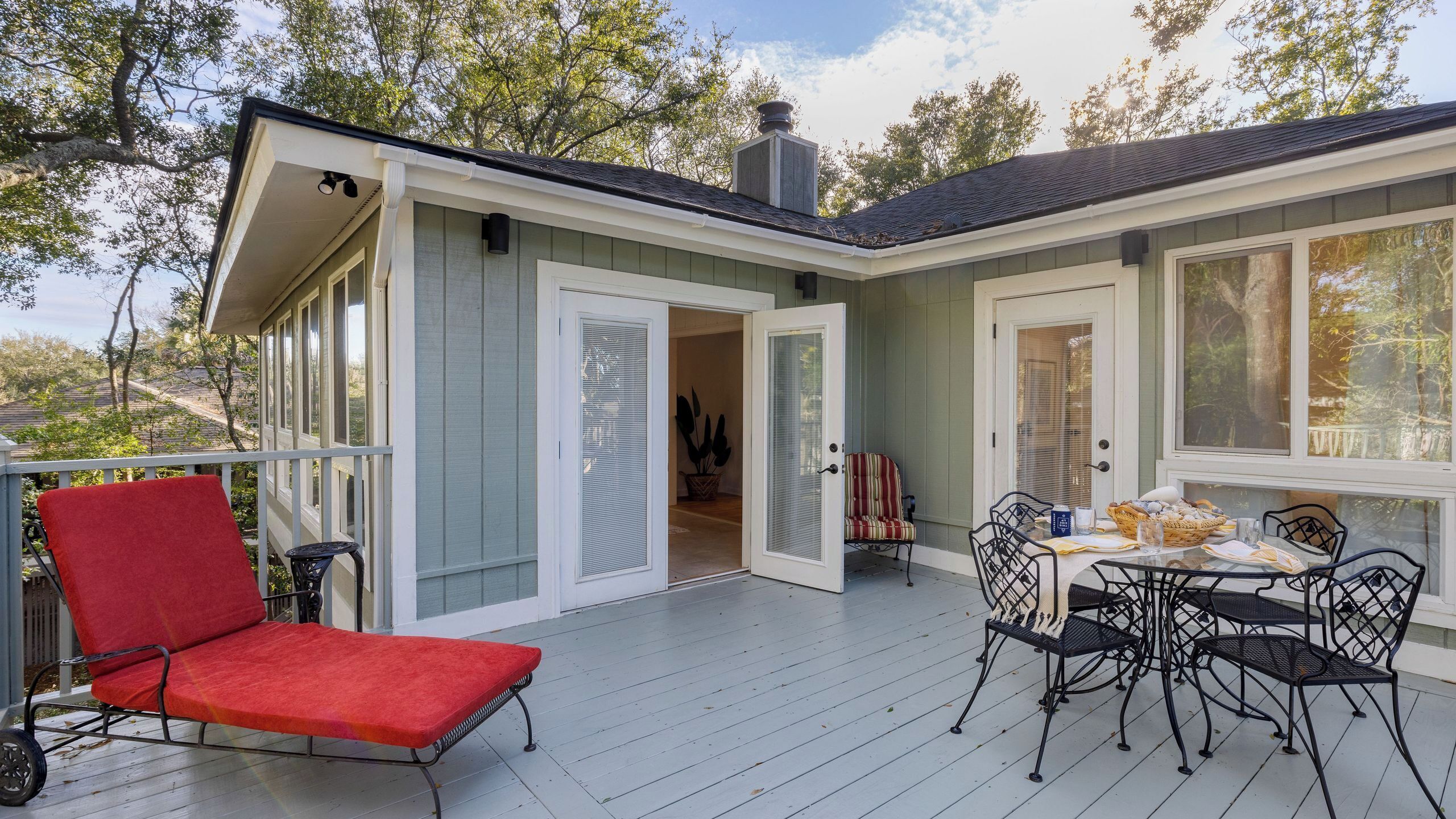 A covered deck with a red chair, a dining table with chairs, and a door leading to an interior room.