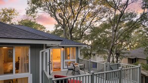 A two-story house with a balcony, a covered outdoor seating area, and a visible bedroom with a bed and curtains. - Conch Cottage by Avantstay Walk to Beach, Deck, Fireplace, Outdoor Dining (Isle of Palms)