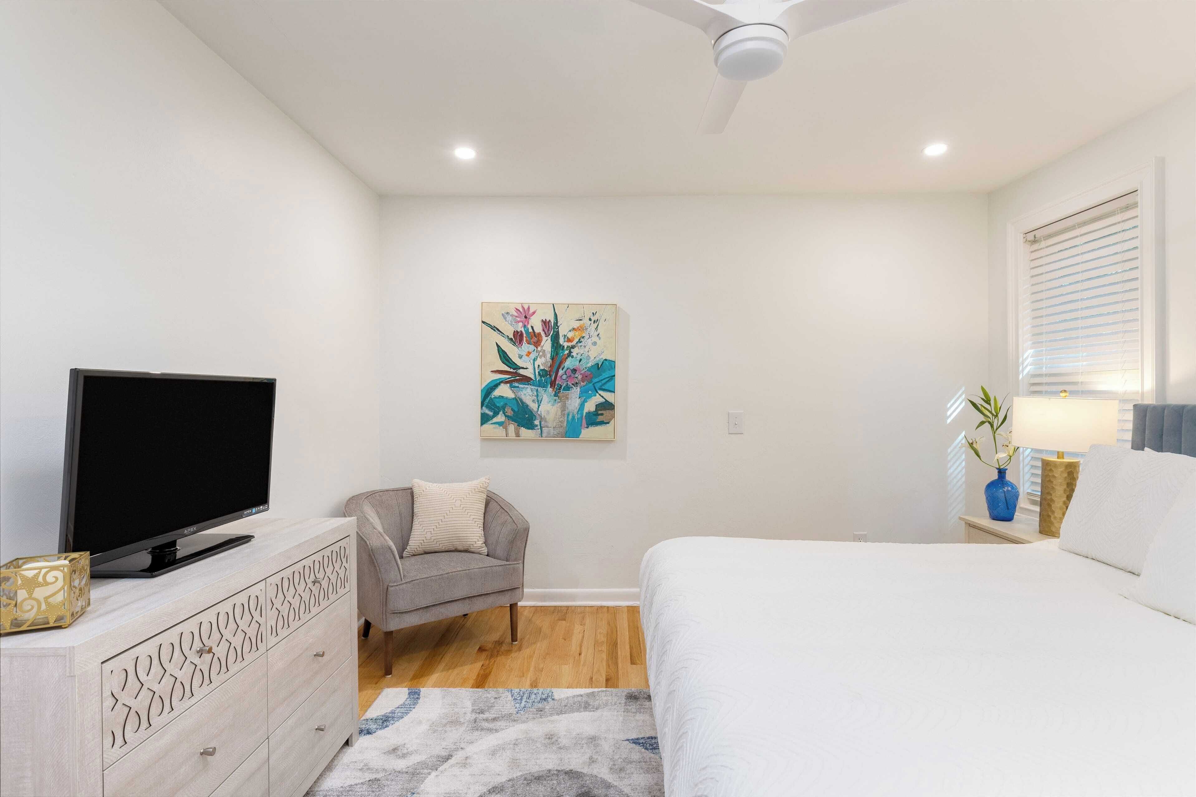 Une chambre à coucher avec un lit, un téléviseur posé sur une commode, un canapé gris, un tableau accroché au mur et un ventilateur de plafond.