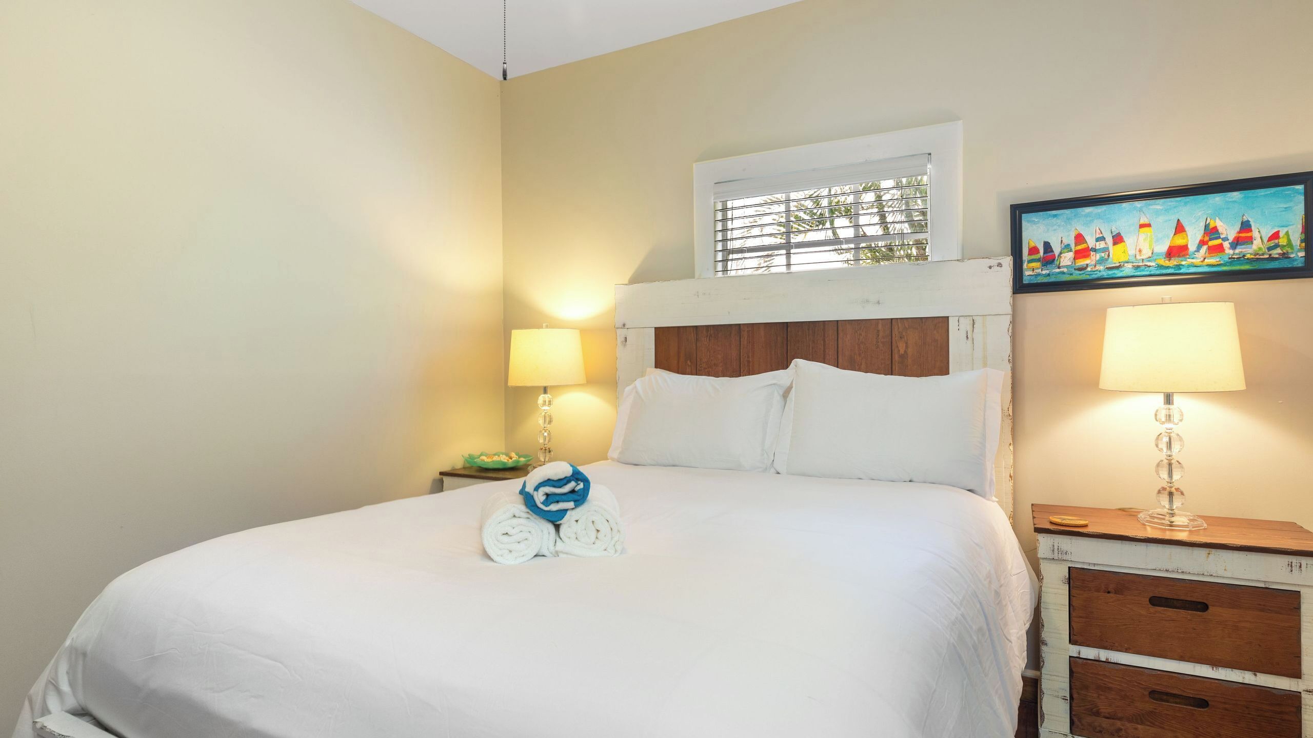 A neatly made bed with white linens, a wooden headboard, a window with blinds, a painting of sailboats, and a bedside lamp.