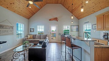 A spacious living area with a high ceiling, wooden beams, a large window, a sofa, a coffee table, and a kitchen with granite countertops.